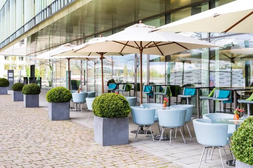 Outdoor seating and parasols beside the glass frontage of Novotel Munich Messe. Photo via DTD 2026's official venue page.